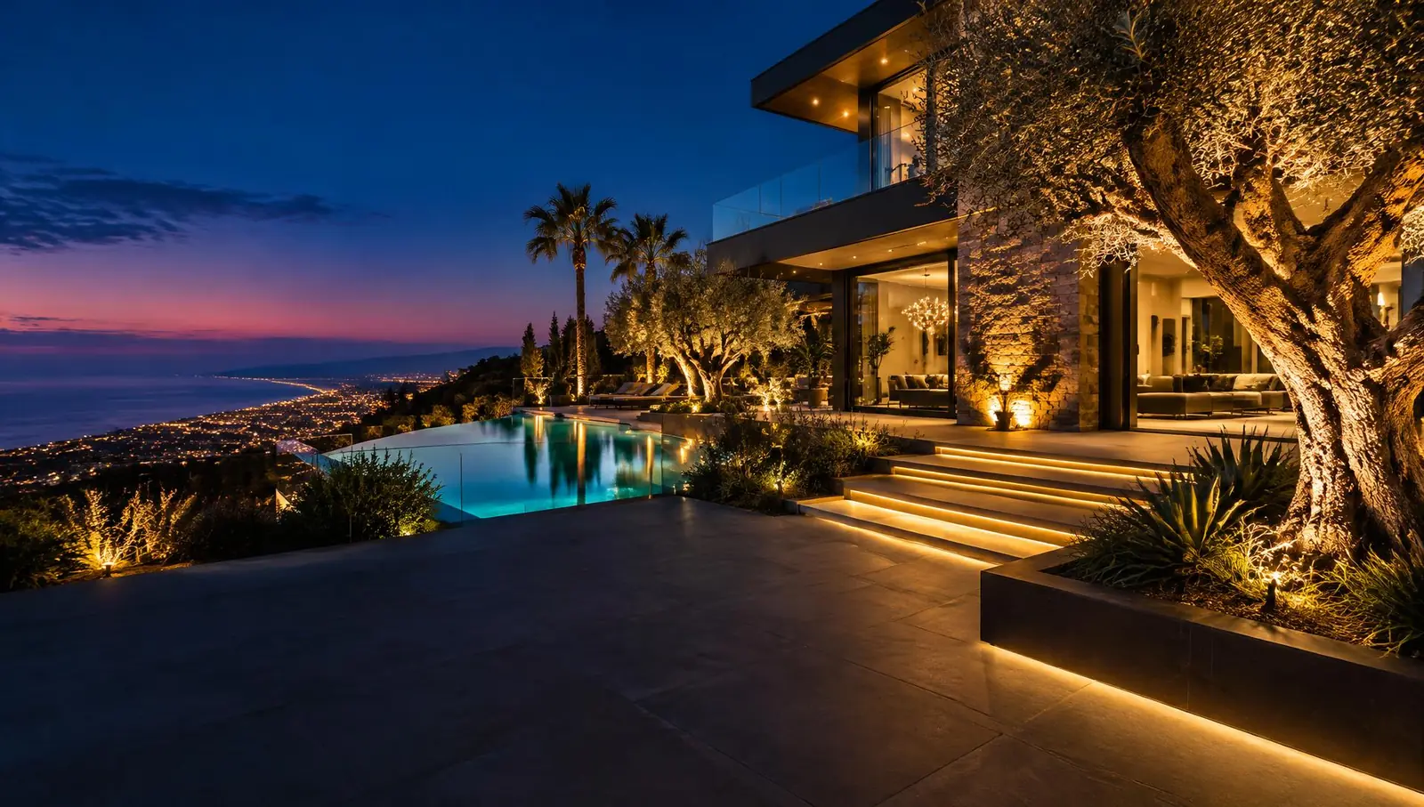 Modern hillside home at dusk with warm landscape lighting, uplit olive tree and glowing infinity pool